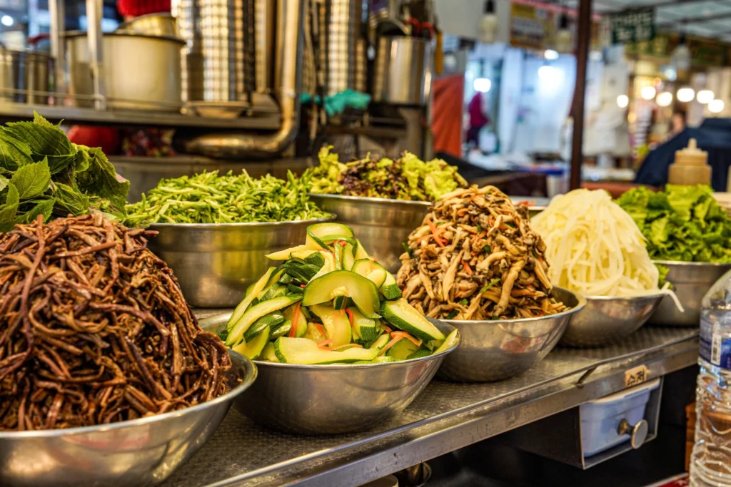 A row of stainless steel bowls filled with individually seasoned namul toppings at Gabose Bibimbap, including gosari bracken fern, zucchini, mushrooms, perilla leaves, and shredded cabbage