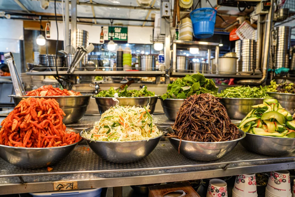 Wide view of the Gabose Bibimbap stall counter showing multiple bowls of colourful namul toppings including spicy radish, cabbage, gosari bracken fern, and cucumber, with the 쌀 보리밥 sign visible in the background