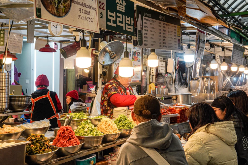 Wide shot of Gabose Bibimbap Stall A22 at Gwangjang Market showing the 쌀 보리밥 sign, the owner preparing food, customers seated at the counter, and colourful vegetable toppings lining the stall front