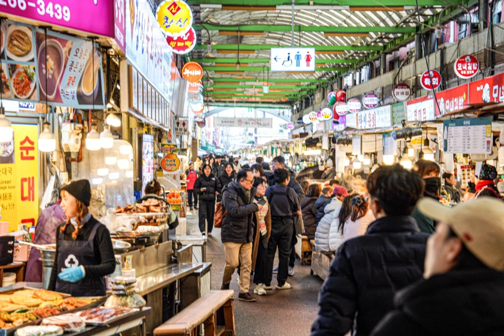 The bustling covered food alley of Gwangjang Market in Seoul, lined with food stalls on both sides and crowded with local diners and tourists