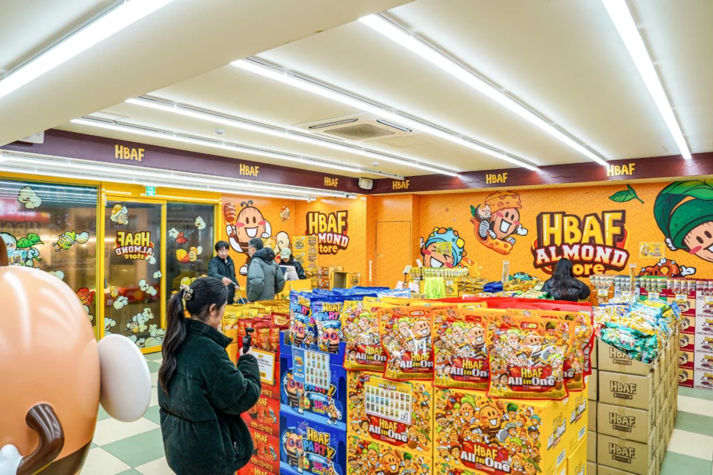 Wide interior view of the HBAF Almond Store at Gwangjang Market, showing orange cartoon-themed walls, product displays, and visitors browsing Korean flavored almond snacks.
