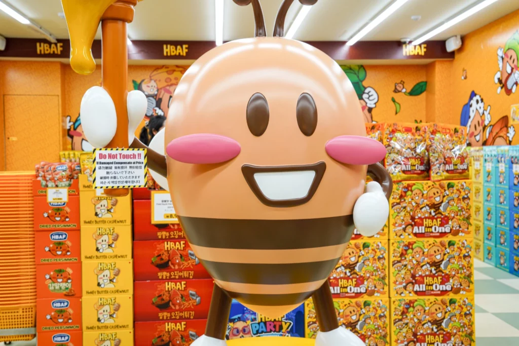 HBAF bee mascot statue smiling at the Gwangjang Market almond store, surrounded by stacked product boxes including Honey Butter Cashew and All in One almond variety packs.