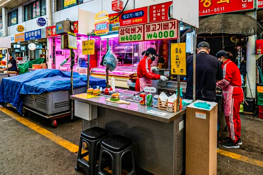 Full exterior view of Samdeok Burger stall at Anyang Jungang Market with staff in red uniforms preparing bulgogi burgers, price boards showing 4,000 won per item.