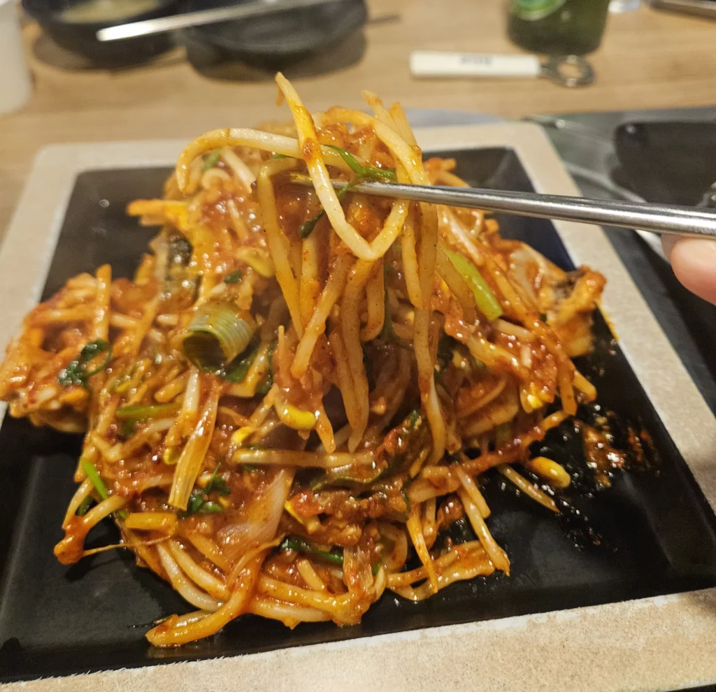 Agujjim: Garosu-gil Braised Monkfish with spicy sauce and bean sprouts being lifted with chopsticks, showing tender monkfish and vegetables on a black plate at a Korean seafood restaurant.