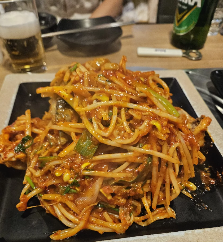 Agujjim: Garosu-gil Braised Monkfish served on a black plate, featuring spicy red sauce, bean sprouts, green onions, and tender monkfish pieces, photographed at a Korean restaurant table with beer in the background.