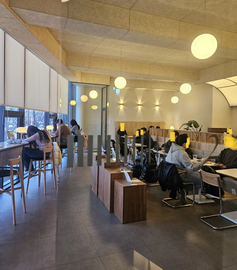 The warmly lit interior of Gabaedo café near Seoul City Hall features guests seated at a mix of window-side bar stools and lounge seating, beneath a geometric textured ceiling with glowing orb pendant lights and soft wall sconces, creating a cozy and stylish atmosphere.