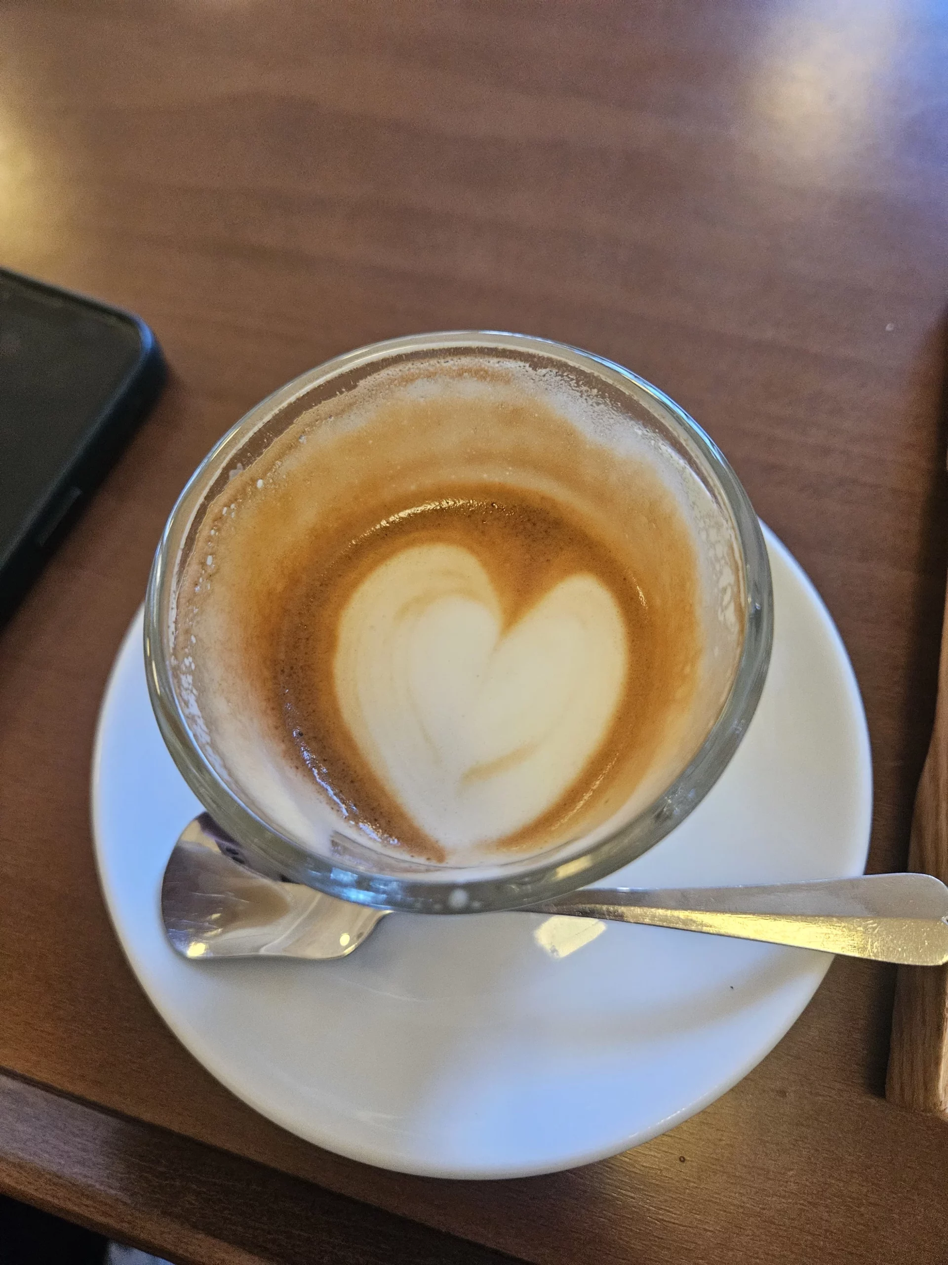 A overhead view of a latte served in a clear glass cup at Gabaedo café near Seoul City Hall, featuring a delicate heart-shaped latte art on a white saucer with a spoon on a wooden table.