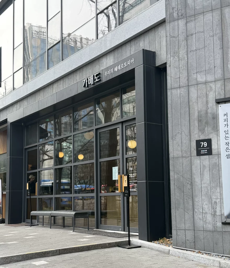 Exterior of Gabaedo (가배도) café near Seoul City Hall, featuring a sleek modern facade with dark steel-framed floor-to-ceiling windows, warm pendant lighting visible inside, and a Korean sign reading 