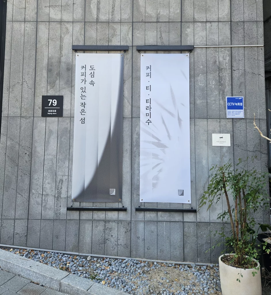 Exterior stone wall of Gabaedo café at 79 Sejong-daero near Seoul City Hall, featuring two elegant vertical hanging banners with Korean calligraphy-style typography. The left banner reads 