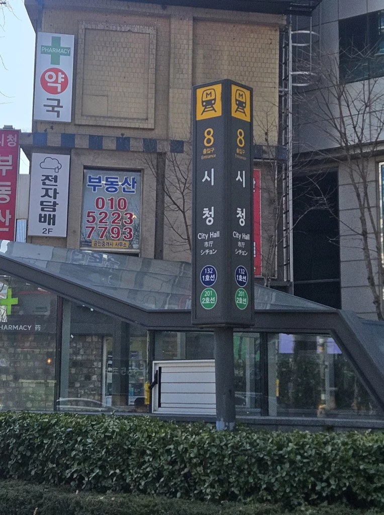 A Seoul Metro station entrance sign for 시청 (City Hall) Exit 8, serving subway Lines 1 and 2, located steps from Gabaedo café, with a glass-covered stairway entrance and street-level shops visible in the background.