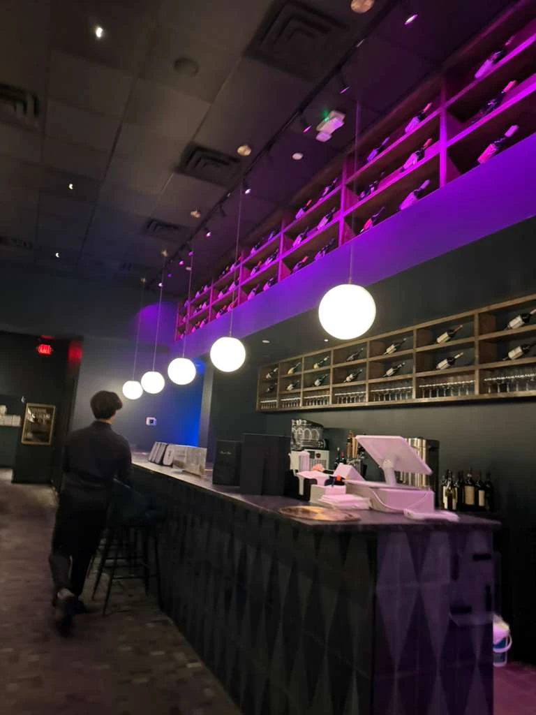 Seoul Prime Korean steakhouse bar area with purple ambient lighting, white globe pendant lights, floor-to-ceiling wine bottle shelving, and a solo diner seated at the bar counter with built-in tabletop grills.
