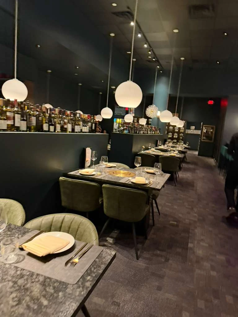 Seoul Prime Korean steakhouse dining room interior with deep teal walls, white globe pendant lights, green velvet chairs, marble tabletops with built-in brass grills, and a curated wine bottle display along the wall.