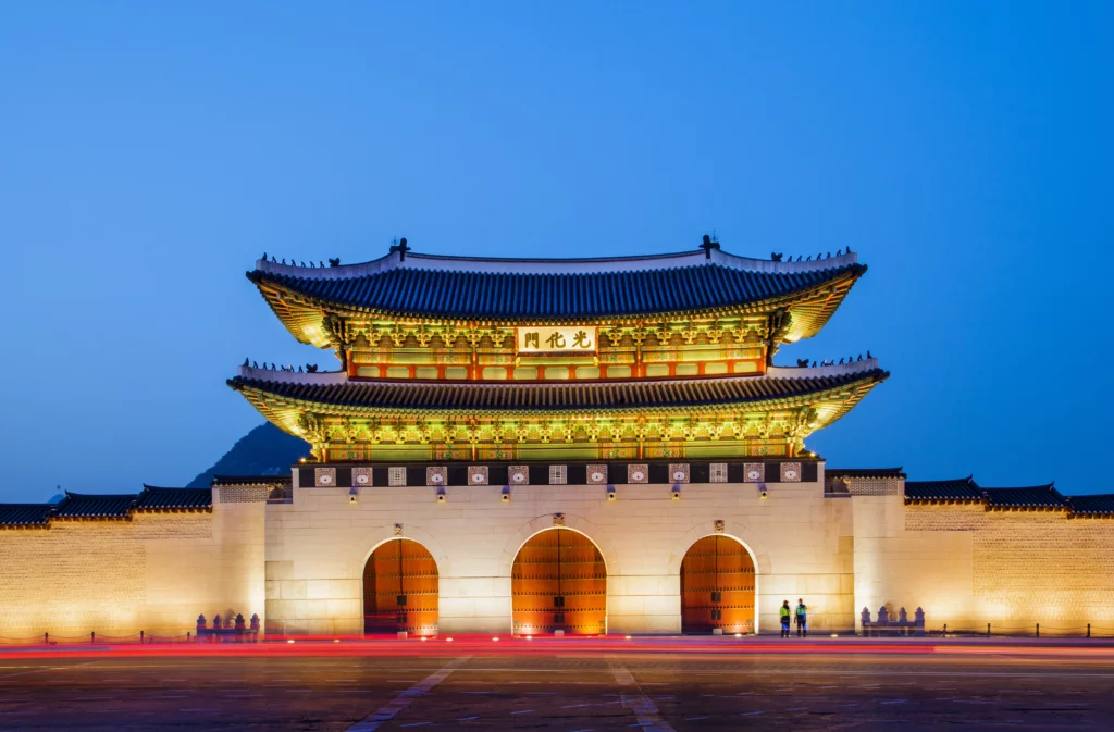 Gwanghwamun Gate (광화문), the grand main entrance of Gyeongbokgung Palace in Seoul, illuminated in vibrant gold and green traditional dancheong colors against a deep blue twilight sky, with light trails from passing traffic in the foreground — a historic landmark within walking distance of Gabaedo café near City Hall.
