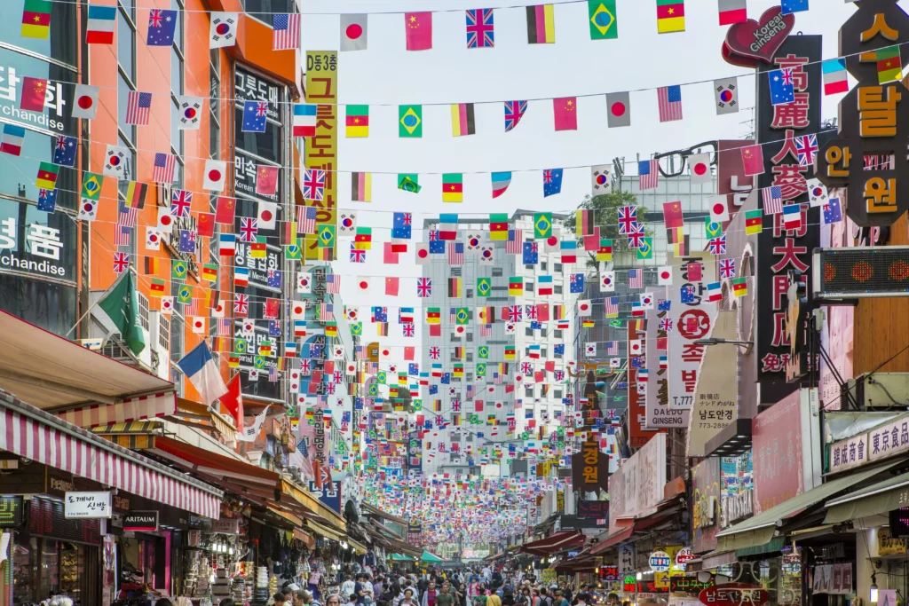 A bustling street at Namdaemun Market in Seoul, canopied with hundreds of colorful international flags strung overhead, flanked by densely packed shops with Korean and multilingual signage, drawing crowds of shoppers — a vibrant landmark near Gabaedo café at City Hall.