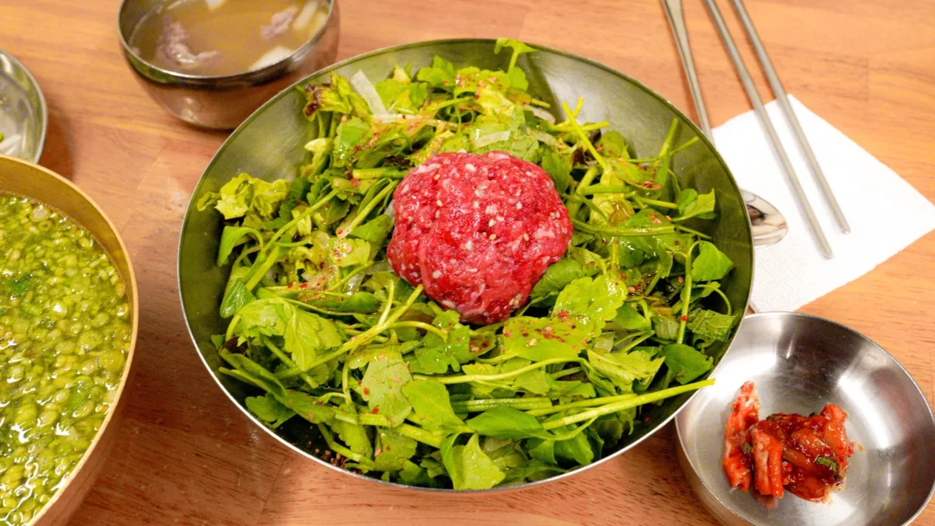 Neungdong Minari fresh Korean water parsley salad topped with raw beef (yukhoe) and sesame seeds served in a metal bowl with dipping sauces.