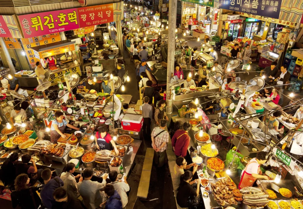 Overhead view of Gwangjang Market's bustling Korean street food alley packed with vendors, diners, and glowing stall lights.