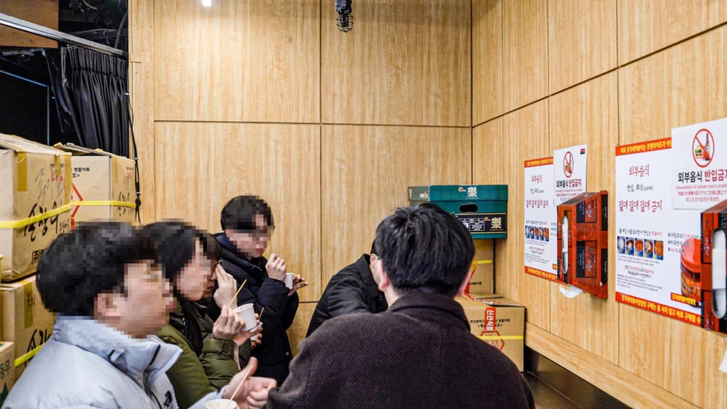 Customers eating tteokbokki while standing at the back counter of Gangga-ne Tteokbokki, with no-outside-food prohibition signs posted on the wooden wall behind them.
