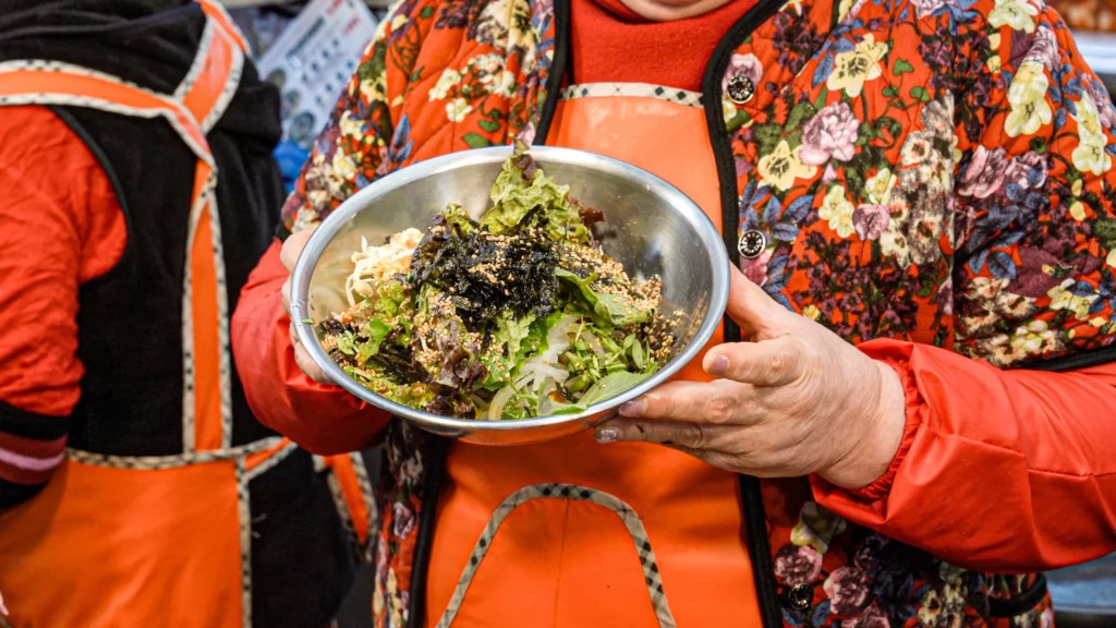 Gabose Bibimbap owner holding a freshly assembled barley bibimbap bowl topped with mixed greens, dried seaweed, and sesame seeds