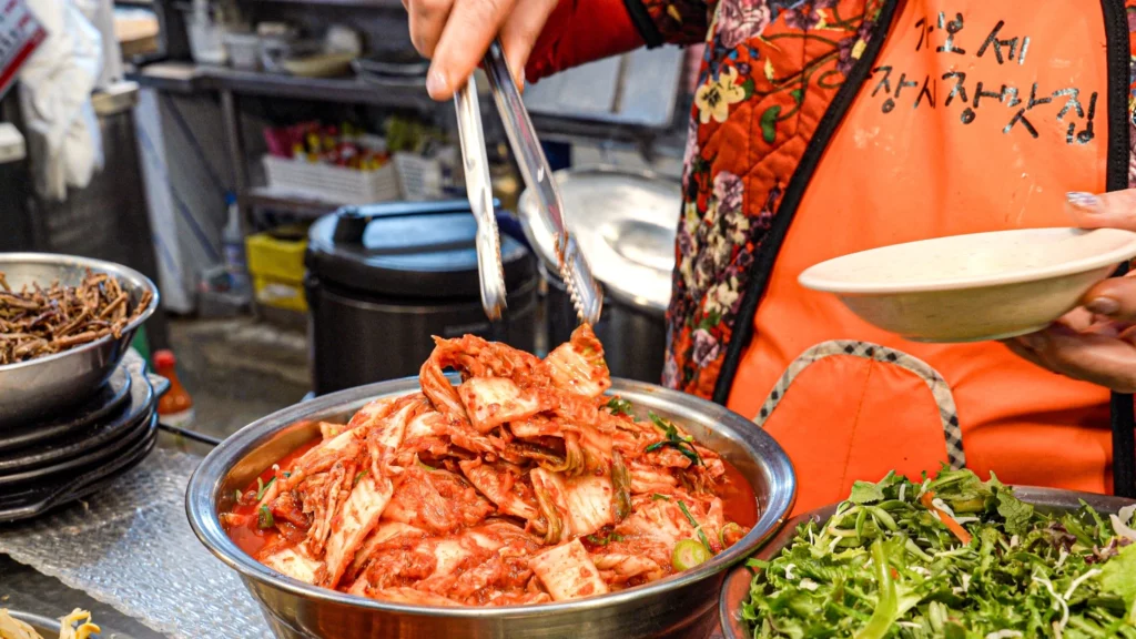 Gabose Bibimbap owner in an orange apron using tongs to serve kimchi, with fresh greens visible in the foreground — the apron reads Gabose Gwangjang Market