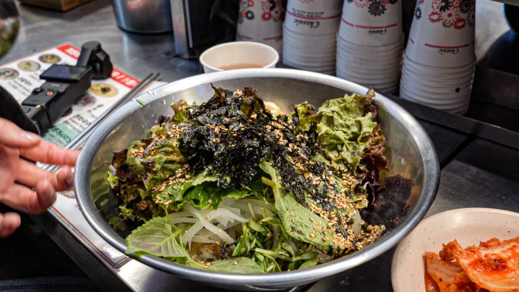 A stainless steel bowl of barley bibimbap at Gabose Bibimbap, topped with fresh mixed greens, crispy seaweed flakes, and toasted sesame seeds, with kimchi served on the side