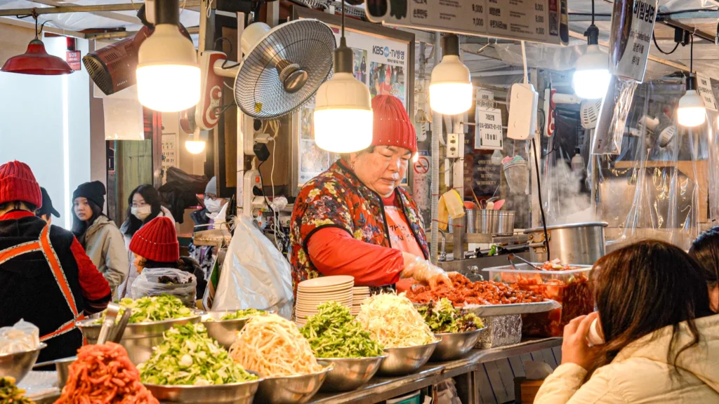 The owner of Gabose Bibimbap at Gwangjang Market prepares fresh vegetable toppings behind the counter, surrounded by large bowls of namul ingredients