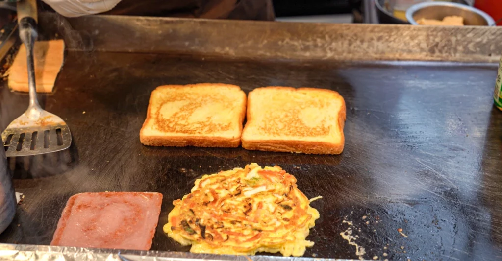 Golden butter-griddled bread slices, a vegetable egg omelet, and ham cooking on the Gwangjang Toast hot griddle.