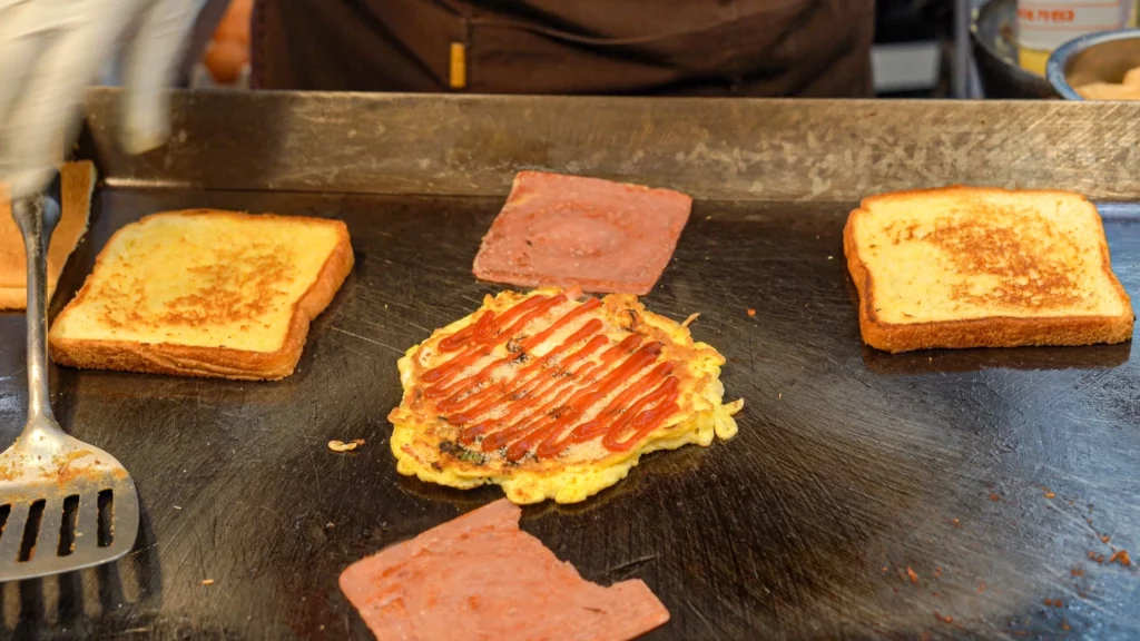 Vendor drizzling ketchup over a fried egg omelet on the griddle with butter-toasted bread and ham slices during Korean street toast preparation.
