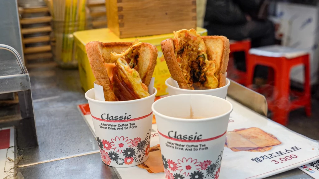 Two freshly made Gwangjang Toast Korean street toast sandwiches cut diagonally and served upright in paper cups at the stall.