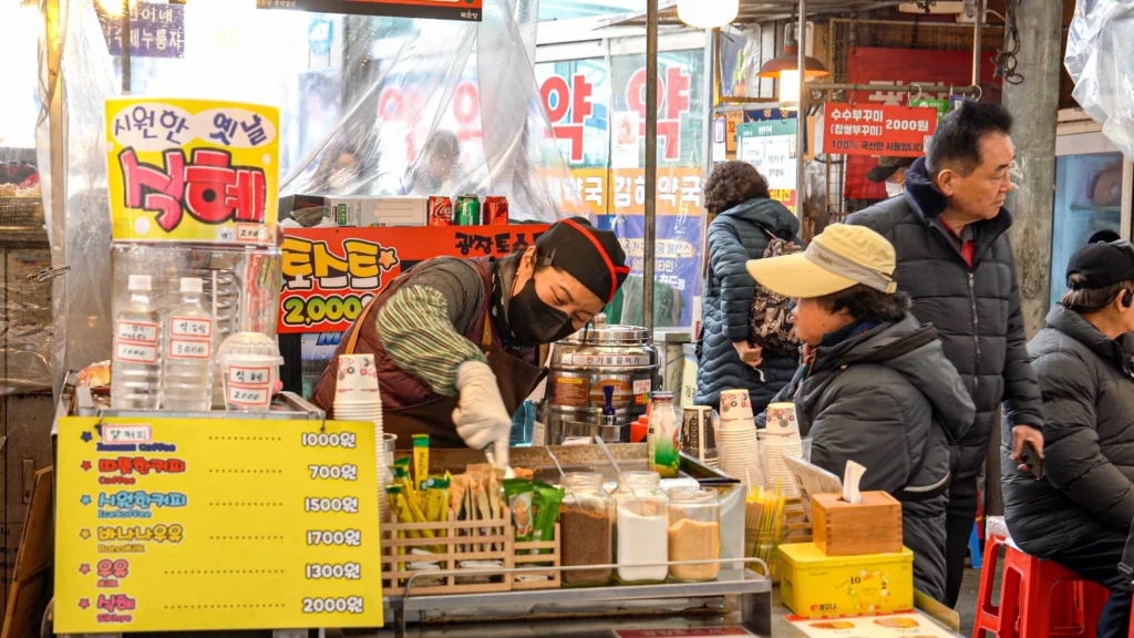 Gwangjang Toast vendor preparing Korean street toast at Dongbu B Stall No. 24 with drink menu board showing prices from ₩700 to ₩2,000.
