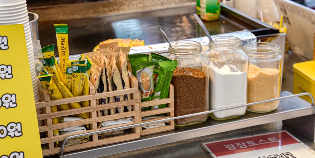 Maxim instant coffee stick packets, tea sachets, and jars of sugar and creamer lined up at the Gwangjang Toast counter labeled 