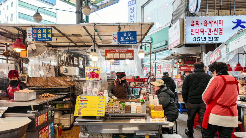 Wide view of Gwangjang Toast at Dongbu B Stall No. 24 inside Gwangjang Market's food corridor with vendor, customers, and neighboring bindaetteok stall