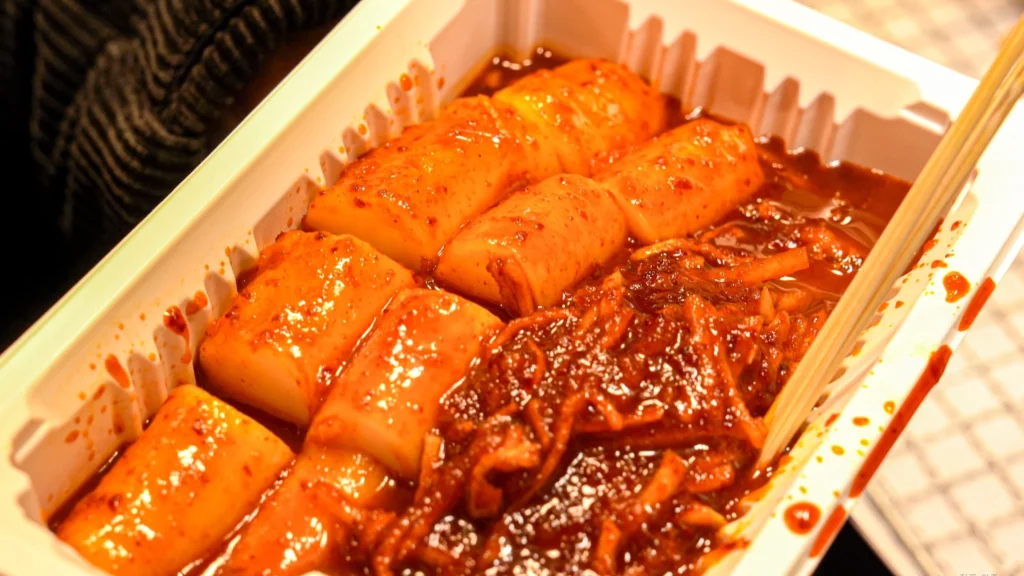 Gangga-ne mu-chae tteokbokki served in a white styrofoam container: thick cylindrical rice cakes in deep-red radish-gochujang sauce with shredded radish strands visible.<br>
