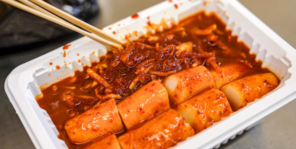 Close-up of Gangga-ne mu-chae tteokbokki in a white styrofoam container, chopsticks lifting a cylindrical rice cake coated in thick radish-gochujang sauce.