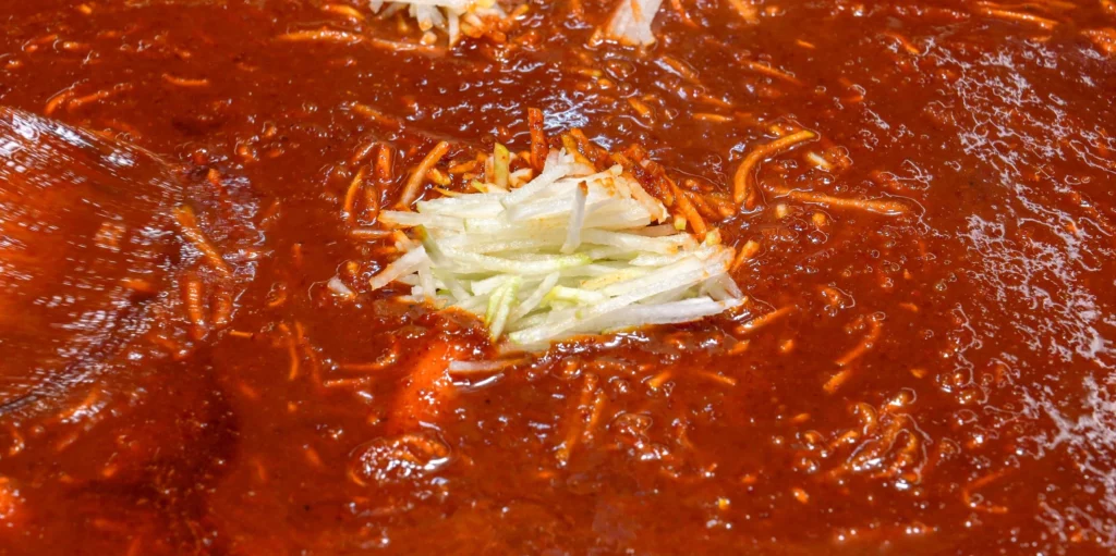 Extreme close-up of Gangga-ne Tteokbokki's boiling gochujang broth with shredded radish strands dispersed throughout, showing the distinctive mu-chae sauce texture.