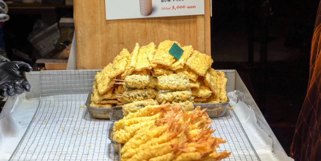 Freshly fried twigim Korean street food fried snacks including tempura-battered vegetables and seafood piled on a wire mesh rack, served as a pairing option with Gangga-ne tteokbokki.<br>