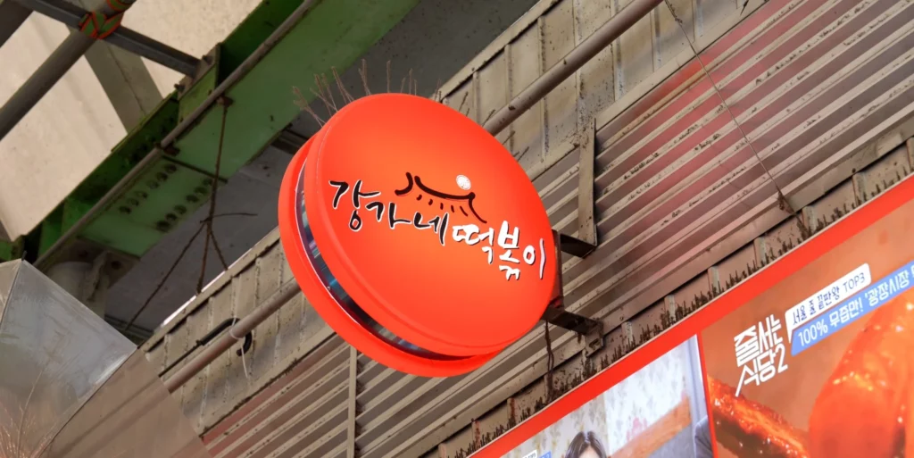Round orange hanging sign with Gangga-ne Tteokbokki in Korean calligraphy suspended from the Gwangjang Market ceiling, marking the stall's location near the entrance.