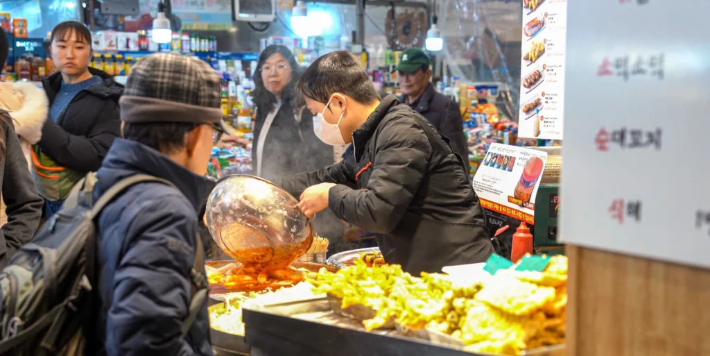 Gangga-ne Tteokbokki vendor serving a customer by ladling tteokbokki sauce over an order, with freshly fried twigim laid out on the counter and a queue of customers waiting in the steam-filled stall.<br>