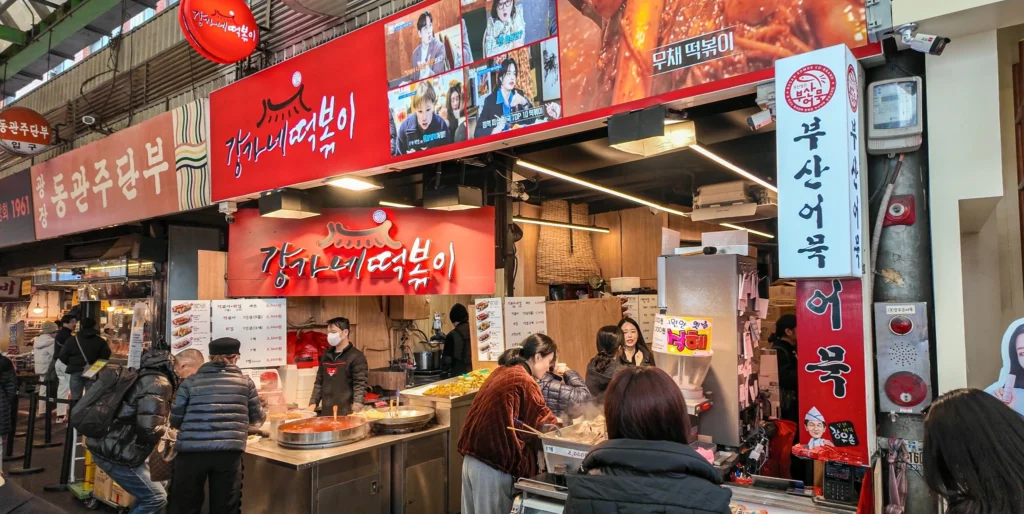 Full exterior view of Gangga-ne Tteokbokki stall inside Gwangjang Market, with red signage reading Gangga-ne Tteokbokki and Mu-chae Tteokbokki, steaming pots, and customers eating while standing.<br>