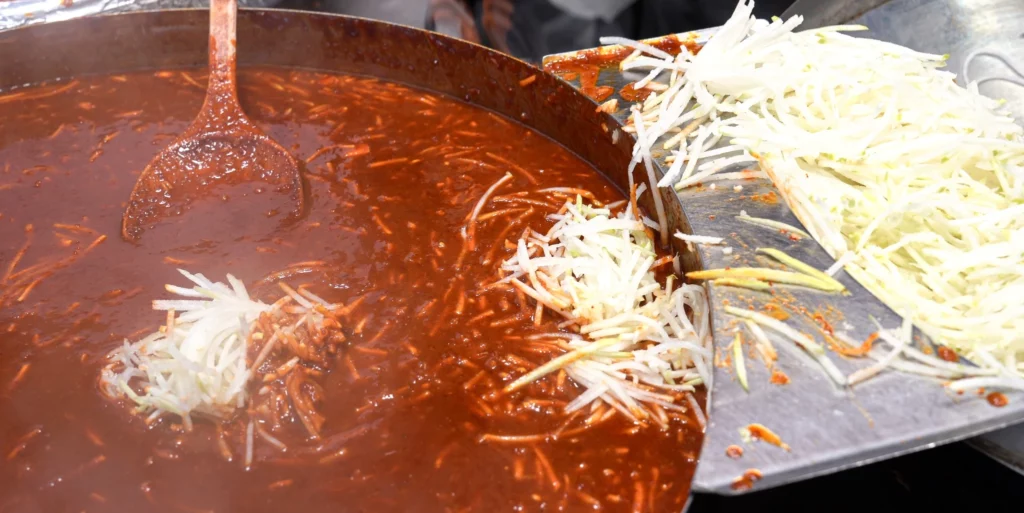 Shredded Korean radish being added in large quantities to a wide flat wok of deep-red gochujang sauce — Gangga-ne Tteokbokki's signature radish-broth technique