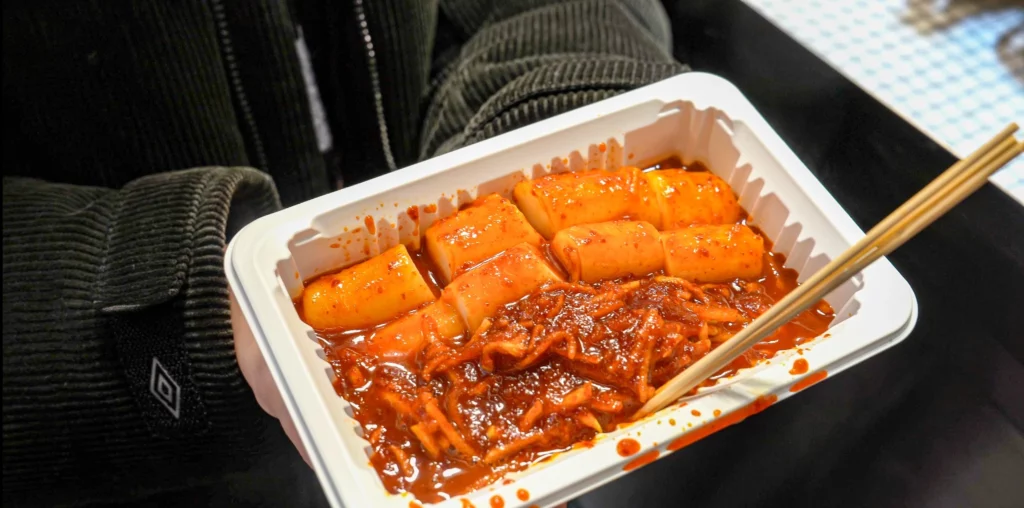 Customer holding a styrofoam container of Gangga-ne mu-chae tteokbokki with chopsticks while eating standing — the traditional pojangmacha-style street food experience.<br>
