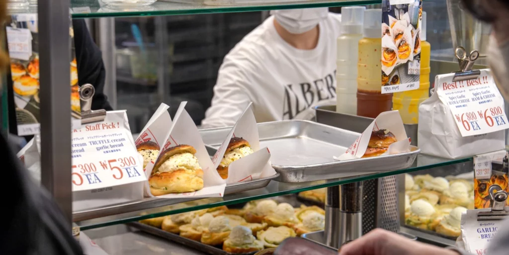 Garlic Boy display counter showing truffle cream garlic bread at ₩5,300 and spicy garlic ham & cheese bread at ₩6,800, with a staff member serving customers.