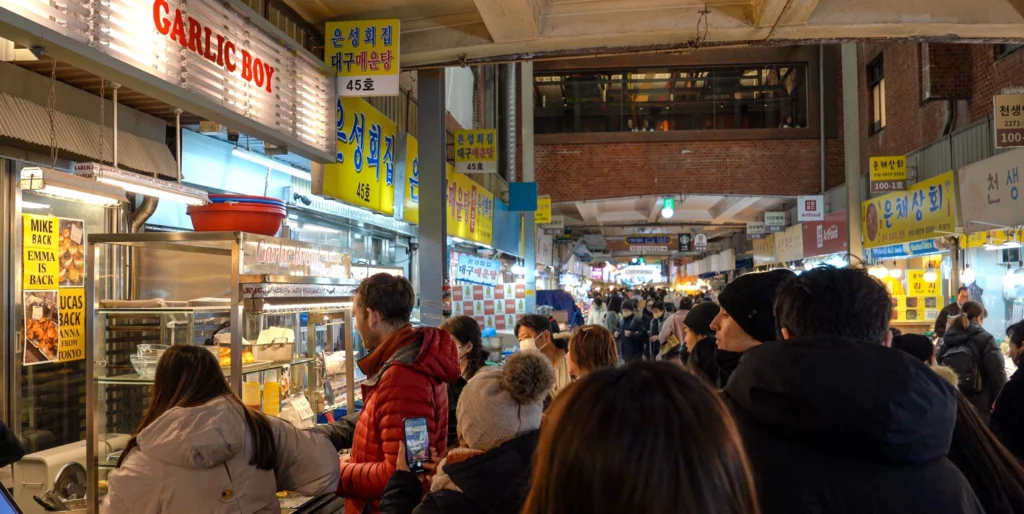 A long queue of customers waiting to order at the Garlic Boy stall inside Gwangjang Market, with the lit 