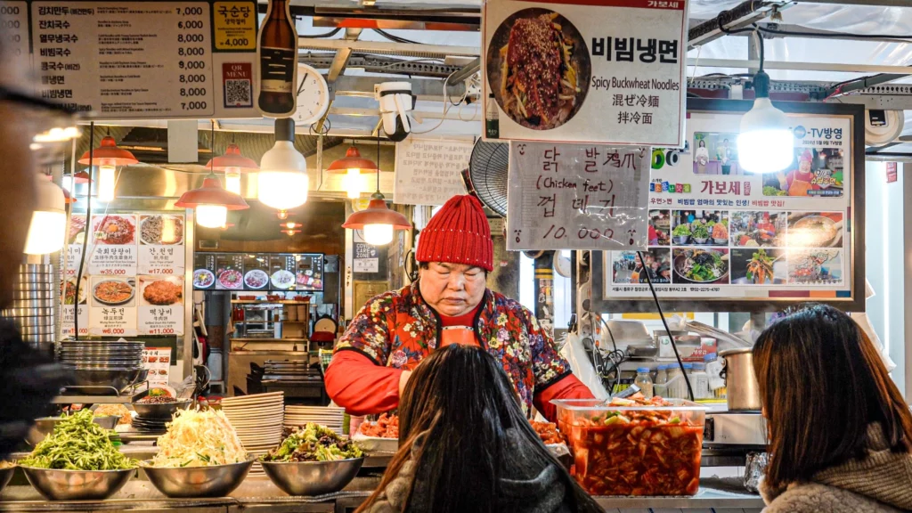 Front view of Gabose Bibimbap Stall A22 at Gwangjang Market with the owner serving customers, bibim naengmyeon banner overhead, and a KBS TV feature poster visible on the right