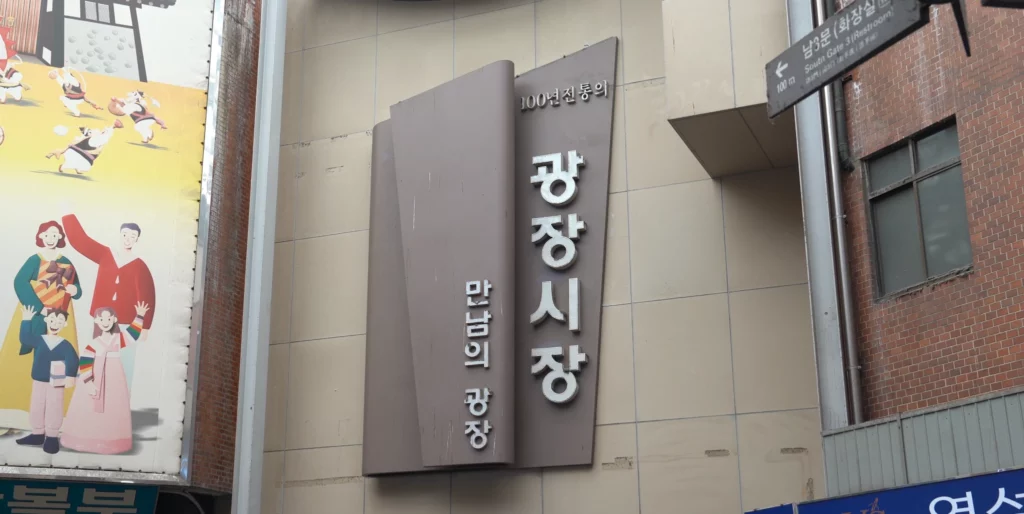 Exterior entrance sign of Gwangjang Market reading 100 Years of Tradition, with a traditional Korean illustration mural visible beside it.<br>