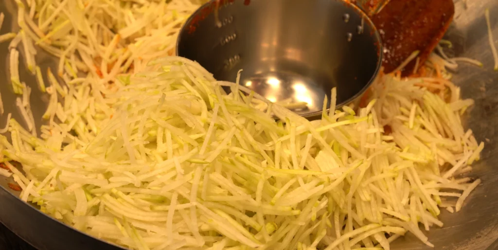 Large bowl of finely shredded Korean radish with gochujang sauce being added by ladle, showing the key ingredient preparation for Gangga-ne's radish-based tteokbokki.<br>