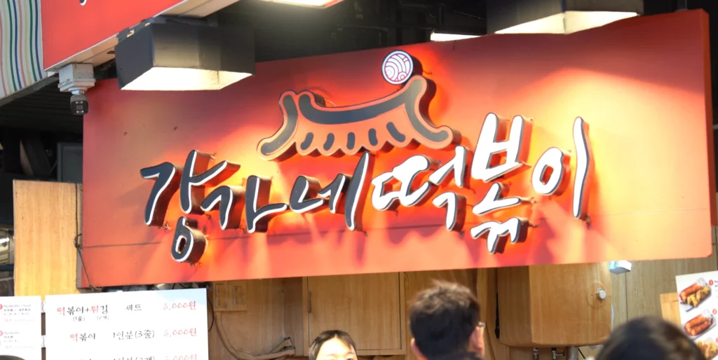 Illuminated red signboard reading Gangga-ne Tteokbokki above the stall entrance, with the price menu board below showing tteokbokki-and-fried set and single-serving tteokbokki both at 6,000won.