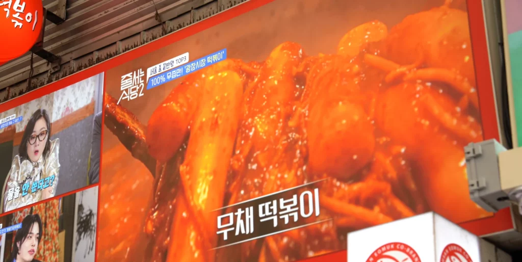 Outdoor digital display screen at Gangga-ne Tteokbokki showing a Korean TV program featuring the stall as one of Seoul's top three queue-worthy tteokbokki spots, with a close-up of radish tteokbokki on screen.