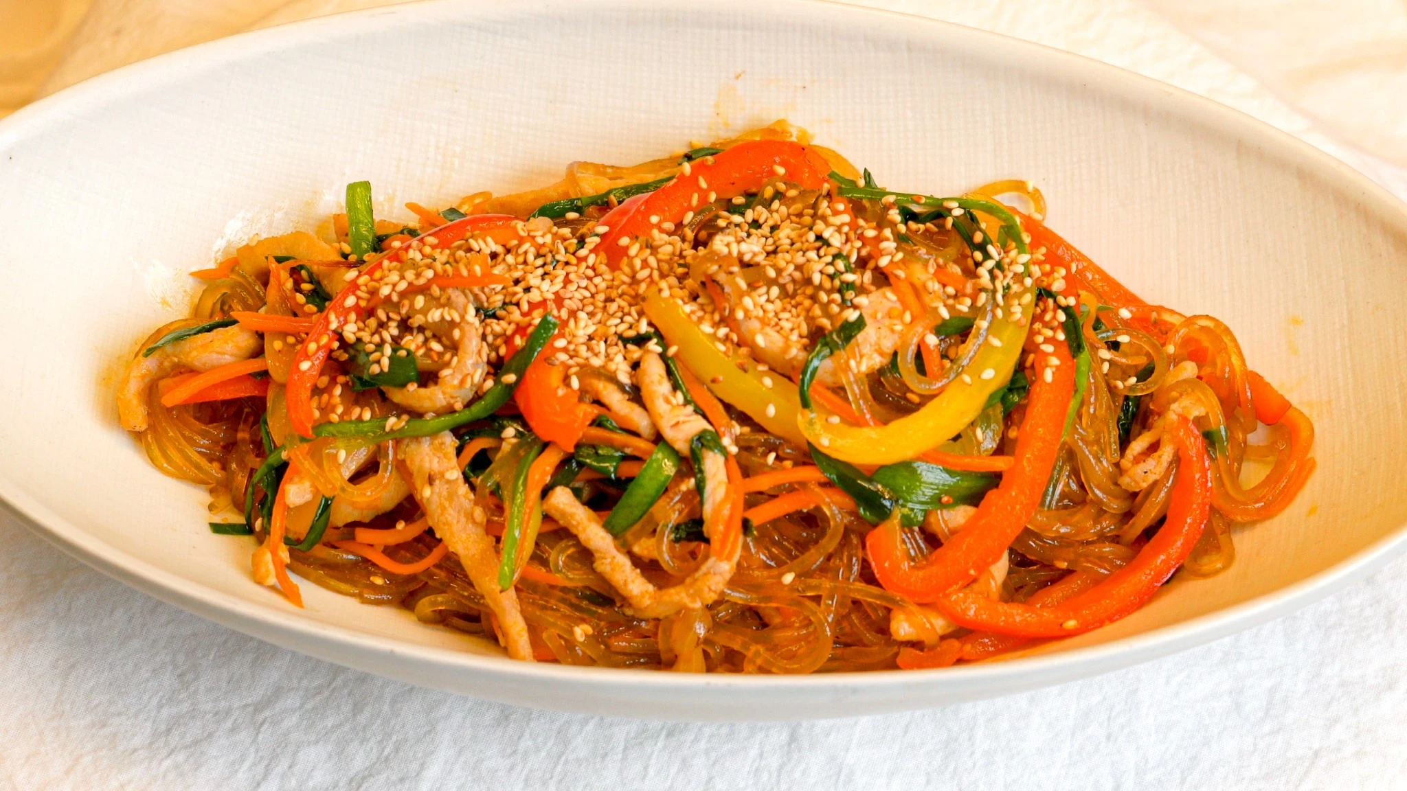 Finished spicy smoky japchae served on a white oval plate, with glossy dark dangmyeon glass noodles topped with stir-fried pork strips, red and yellow bell peppers, julienned carrot, and garlic chives, generously garnished with toasted sesame seeds.