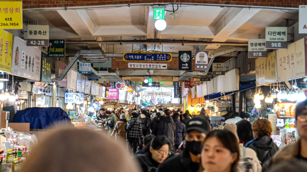 Crowds of visitors filling the busy indoor alley of Gwangjang Market in Seoul, lined with vendor signs on both sides.