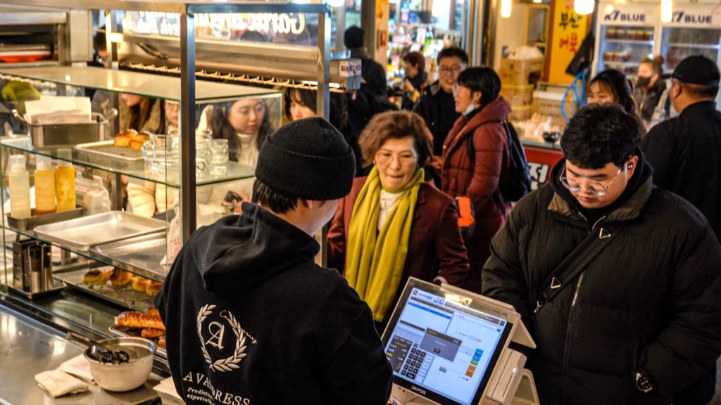 Customers lining up to order at the Garlic Boy stall counter inside Gwangjang Market, with garlic breads visible in the display case.